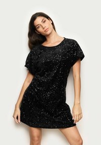 Černé šaty pokryté flitry s krátkými rukávy a volným střihem. Látka má texturovaný, lesklý povrch odrážející světlo.