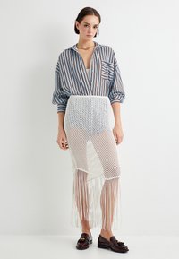 Chemise surdimensionnée rayée bleu et marron, associée à une jupe en filet blanche avec des franges sur l'ourlet. Des mocassins noirs viennent parfaire la tenue.