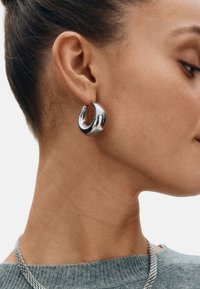 Strieborná náušnica v tvare kruhu s asymetrickým dizajnom, vybavená lešteným a odrážajúcim povrchom a bezpečným zapínaním na šrób.