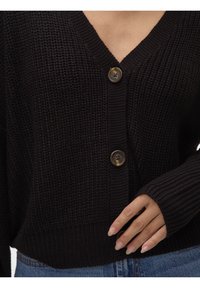 Musta neulottu neuletakki, jossa on V-aukko, resorit hihansuissa ja helmassa sekä kaksi näkyvää ruskeaa nappia edessä. Kankaan tekstuuri lisää syvyyttä.