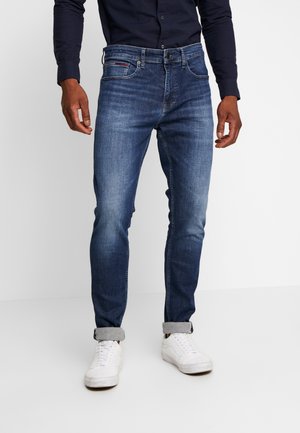 Mężczyzna ubrany w ciemnoniebieskie jeansy typu slim fit z podwiniętymi nogawkami, białe trampki oraz czarną koszulę na guziki z długim rękawem, stojący na białej podłodze.
