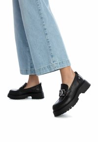 Mocasines de cuero negro con suela gruesa y estriada, detalle de hebilla rectangular y un acabado suave. Combinados con vaqueros de denim azul claro de pierna ancha.