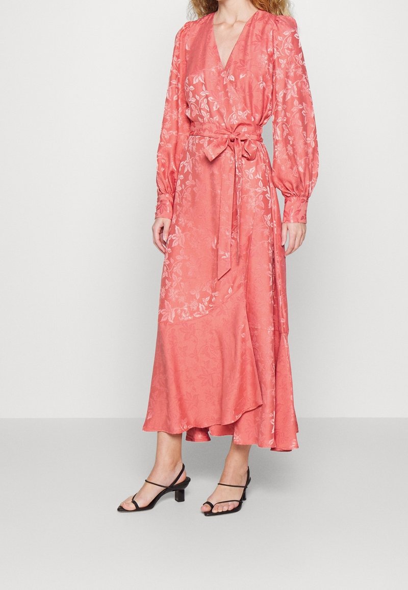 Rosa omlottklänning med långa puffärmar, blommönster, V-ringning, cinchad midja med bälte, och en flödande nederkant, i kombination med svarta sandaler.