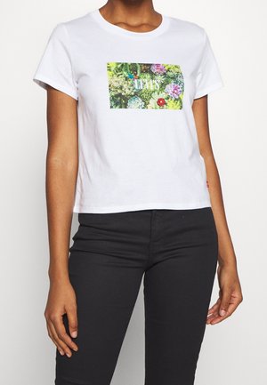 Mujer con camiseta blanca corta con diseño floral colorido y texto "Levi's", combinada con pantalones ajustados negros.