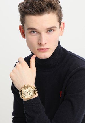 Montre en or avec un cadran rond, des accents noirs et dorés, et un bracelet en métal. Modèle portant un col roulé noir, la main reposant sur le menton.