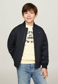 Giubbotto bombardiere navy con polsini elasticizzati, maglione giallo chiaro con testo nero e jeans azzurri. Tessuto morbido e design casual.
