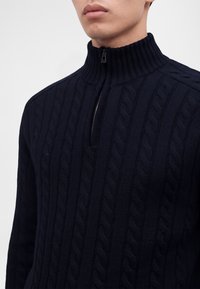 Homme portant un pull en maille torsadée bleu marine foncé avec un col à demi-fermeture éclair partiellement zippé.