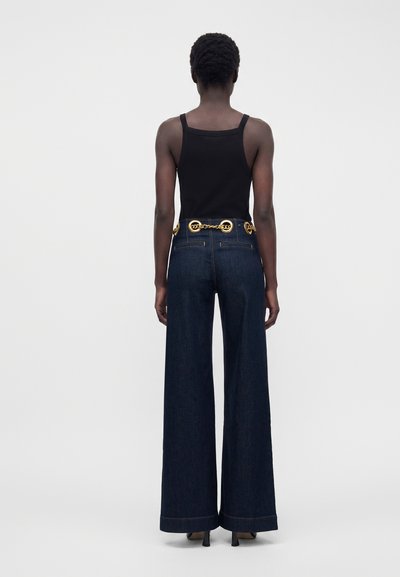 Person iført en sort ærmeløs top, mørke vidde jeans med baglommer og et gyldent bælte med kæde, stående med ryggen mod kameraet på en ensfarvet baggrund.