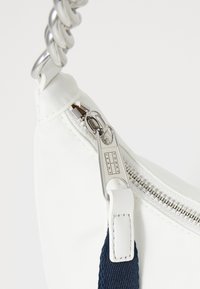 Balta audinė rankinė su sidabriniu užtrauktuku, mėlynu diržo akcentu ir metaliniu grandininiu rankeniu. Tommy Jeans logotipas matomas netoli užtrauktuko.