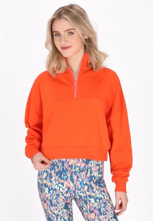 Młoda kobieta o blond włosach nosi jasnopomarańczową bluzę na zamek oraz kolorowe, wzorzyste legginsy, stojąc na jednolitym białym tle.