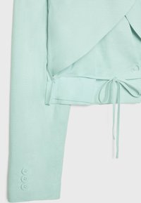 Manche courte bleu clair d'un blazer avec trois boutons et un détail de cordon serreur près de la taille sur fond blanc.