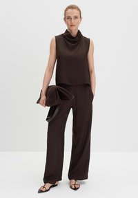 Top marrone senza maniche con scollatura a cowl abbinato a pantaloni marroni larghi. Il modello tiene una borsa strutturata marrone scuro. Tessuto morbido, design minimale.