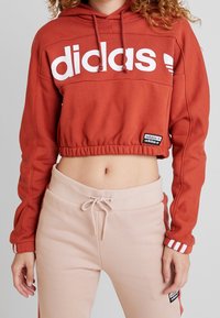 Rostfärgad kort hoodie med vitt "adidas"-logotyp, elastisk midja, känguruficka och ribbade ärmar; matchad med ljusbeiga joggers.