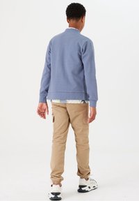 Felpa in cotone blu con una texture a coste, indossata sopra una camicia floreale, abbinata a pantaloni cargo beige e sneakers bianche.