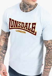 Šviesiai mėlynas medvilninis marškinėliai su apvaliu kaklu, kuriuose didelė bordo spalvos "LONSDALE LONDON" logotipas su ryškiais rašmenimis.