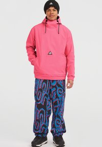 Felle roze pulloverjack met een hoge kraag, koordjes en driehoeklogo; gecombineerd met zwarte broek met blauwe en roze abstracte patronen.