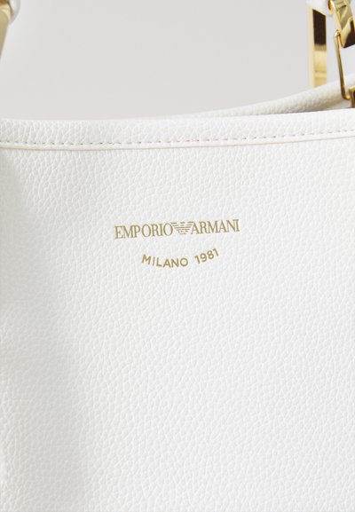 Balta rokassoma ar Emporio Armani zelta logo, zem kura ir iegravēts "MILANO 1981", demonstrējot teksturētu ādas virsmu.