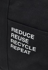 Černá látková taška s bílým textem "REDUCE REUSE RECYCLE REPEAT". Obsahuje přední kapsu s viditelným šitím. Jednoduchý, funkční design.