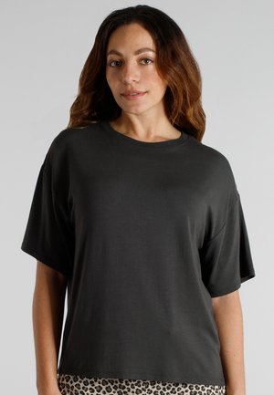 Frau mit welligem braunem Haar, die ein lockeres schwarzes Kurzarmshirt und Leopardenmusterhosen vor einem schlichten grauen Hintergrund trägt.