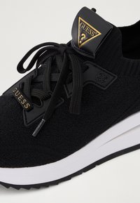 Schwarzer Sneaker mit einem strukturierten Strick-Obermaterial, robusten schwarzen Schnürsenkeln, Kunststoffakzenten und einer weißen Mittelsohle. Verfügt über ein dreieckiges Logo-Patch.