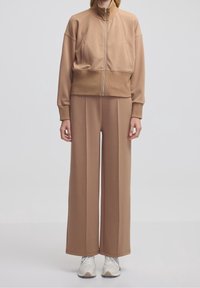 Modella che indossa pantaloni larghi beige abbinati a una giacca cropped con zip, delle sneakers bianche, in piedi contro uno sfondo semplice.