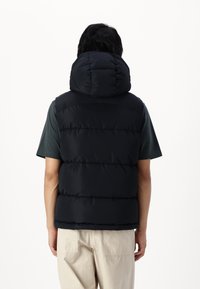 Czarna puchowa kamizelka z wysokim, zaokrąglonym kapturem, z poziomym przeszyciem, zestawiona z krótkim zielonym t-shirtem i jasnymi spodniami.