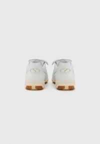 Witte leren sportschoenen met een rubberen zool, voorzien van een geel logo op de hiel en lusjes voor gemakkelijke sluiting.