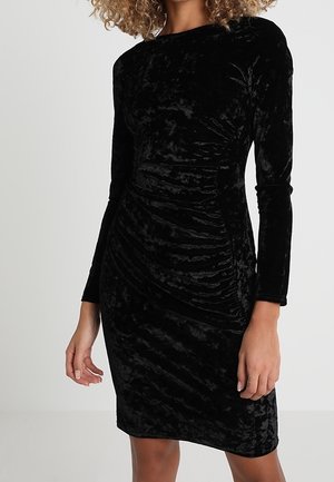 Robe en velours noir avec des manches longues, un décolleté bateau et des détails froncés à l'avant, présentant une silhouette ajustée.