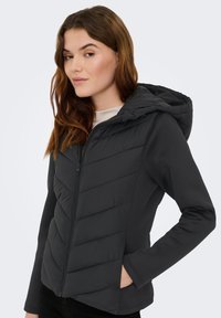 Μαύρο μπουφάν με κουκούλα, με πανέλα με καπιτονέ ραφές και φερμουάρ μπροστά. Μακριά μανίκια και πλαϊνές τσέπες προσθέτουν λειτουργικότητα και ζεστασιά.