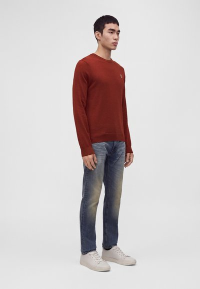 Jeune homme debout portant un pull à manches longues couleur rouille, un jean bleu délavé et des baskets blanches sur un fond clair uni.