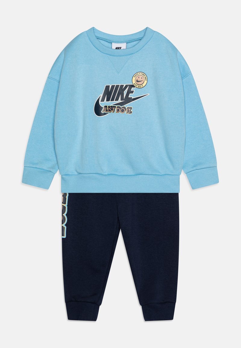 Niebieska bluza z logo Nike i uśmiechniętą buźką; zestawiona z granatowymi spodniami dresowymi z białym napisem po bokach i elastycznymi mankietami.