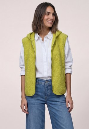 Frau lächelt, trägt eine hellgrüne ärmellose Fleeceweste über einem weißen Hemd mit Knöpfen und blauen Jeans, vor hellem, einfarbigem Hintergrund.