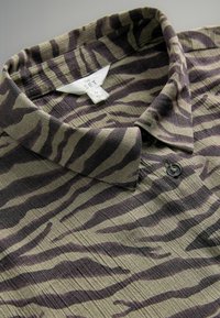 Kariertes Hemd in Khaki mit dunkelbraunen Tigerstreifen, aus strukturierter Stoff gefertigt und mit einem einzelnen schwarzen Knopf am Kragen. Markenetikett sichtbar.