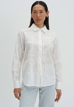 Vrouw met lang donker haar draagt een wit overhemd met knopen en geometrische patronen en lichtblauwe spijkerbroek, staand tegen een eenvoudige achtergrond.