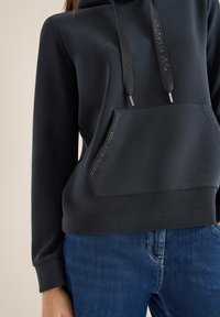 Czarna bluza z kapturem i sznurkiem, z przednią kieszenią ozdobioną małymi kryształkami. Gładka faktura i luźny krój, w połączeniu z niebieskimi dżinsami.