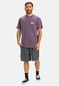 Men's purple cotton t-shirt with a white logo on the left chest, paired with gray cargo shorts. Black socks and black sneakers complete the look.