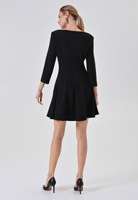 Schwarzes A-Linien-Kleid mit dreiviertellangen Ärmeln, Reißverschluss hinten und glattem Material, kombiniert mit glänzenden schwarzen High Heels.