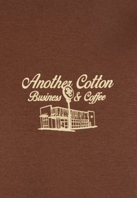 Brun bomullstekstil med hvit brodert tekst: "Another Cotton Business & Coffee" med en linjetegning av en bygning.