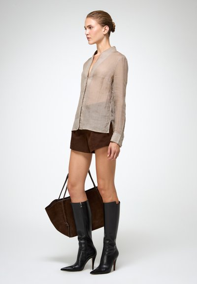 Blusa trasparente beige chiaro, pantaloni corti in suede marrone e stivali alti in pelle nera. Il modello tiene una borsa in suede marrone.