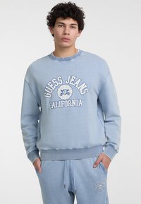 Šviesiai mėlynas apvalaus kaklo megztinis su „GUESS JEANS CALIFORNIA“ baltu tekstu. Minkšta medžiaga su nėriniuotomis rankovėmis ir apačia.