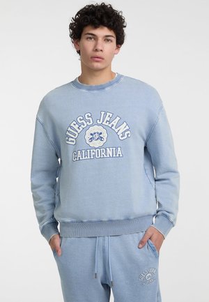 Light blue crewneck sweatshirt featuring "GUESS JEANS CALIFORNIA" in white text. Soft fabric with ribbed cuffs and hem.