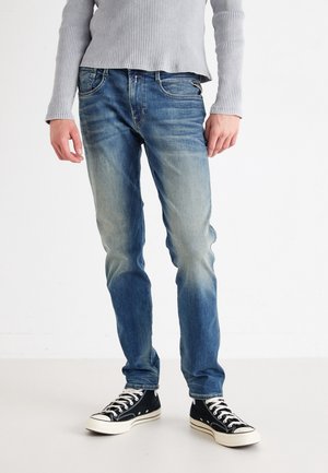 Personne portant un jean bleu délavé, des baskets noires et blanches, et un t-shirt côtelé à manches longues gris clair, debout sur un sol blanc uni.