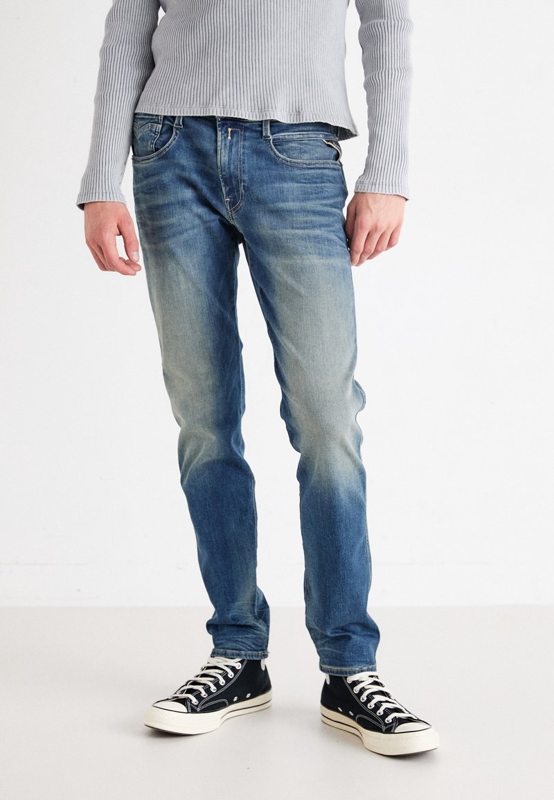 Personne portant un jean bleu délavé, des baskets noires et blanches, et un t-shirt côtelé à manches longues gris clair, debout sur un sol blanc uni.