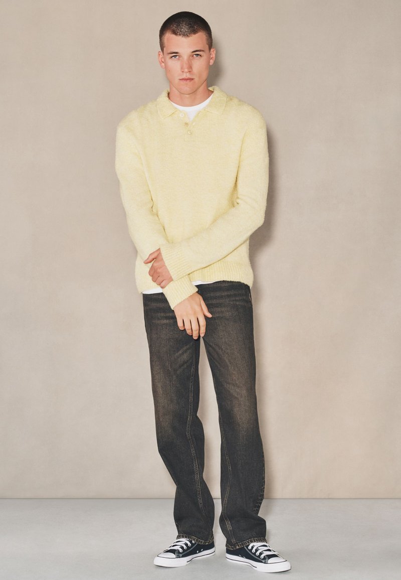 Jeune homme debout portant un pull doux jaune pâle, un jean foncé et des baskets noires basses, posé devant un mur beige uni.