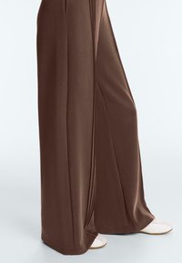 Pantalon marron à jambes larges avec détails sur la couture latérale, porté par-dessus des chaussures blanches sur un fond clair uni.