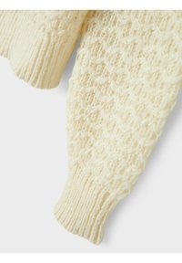 Dettaglio di un maglione lavorato a maglia color avorio con un motivo testurizzato e a trasparenza, e polsini a coste. Il materiale appare morbido e caldo, adatto per le giornate più fresche.