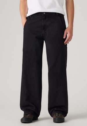 Homme portant un pantalon noir ample à jambes larges avec des poches latérales et des chaussures foncées, debout contre un fond clair uni.