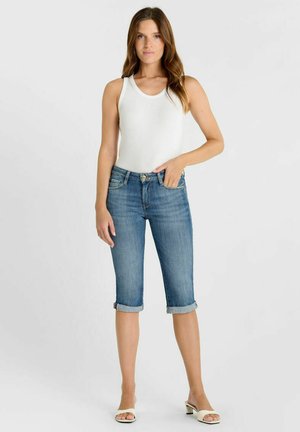 Vrouw die een witte mouwloze top draagt, opgerolde denim capri-jeans en witte open-toe sandalen met hak, staand tegen een effen achtergrond.