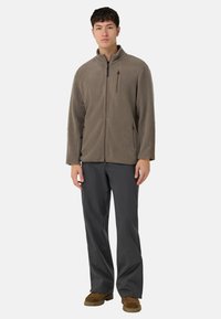 Ruskea fleece-takki, jossa on korkea kaulus, vetoketjullinen rintatasku ja sivutaskut, yhdistetty harmaisiin housuihin ja ruskeisiin kenkiin.