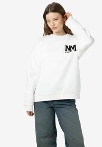 Witte sweater met een losse pasvorm, voorzien van een zwarte geborduurde "NM" logo en de tekst "BE NOISY" op de linkerborst.